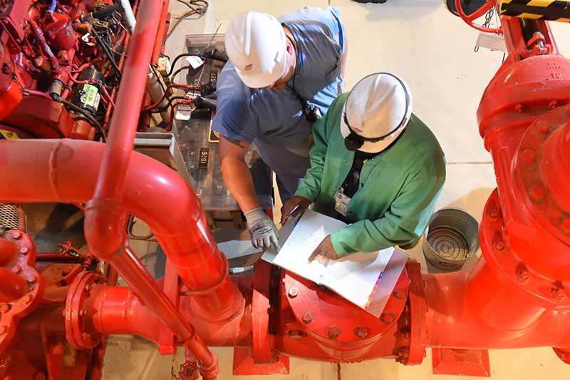 Workers in safety gear reviewing technical plans near large red industrial piping system