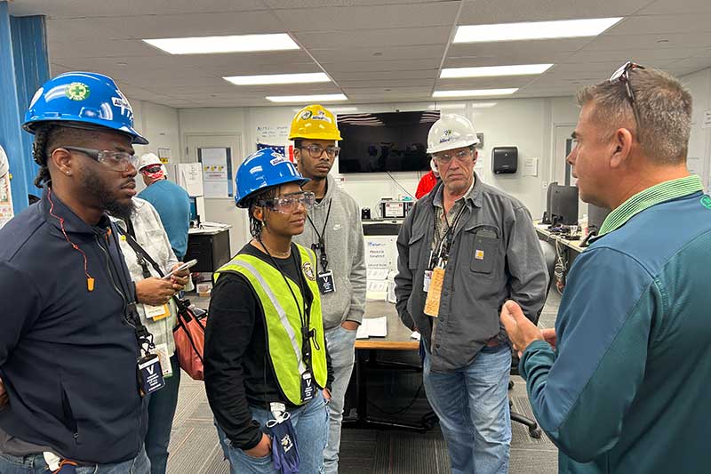Team in hard hats discussing operations inside a control room with monitors and equipment