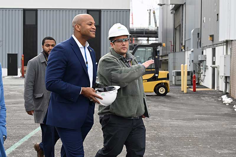 Gov. Wes Moore and workers in safety gear walking outside industrial facility near forklift and large structures